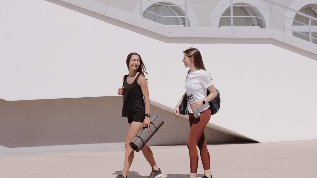 Two women in active wear walking outdoors while smiling and carrying yoga mats.